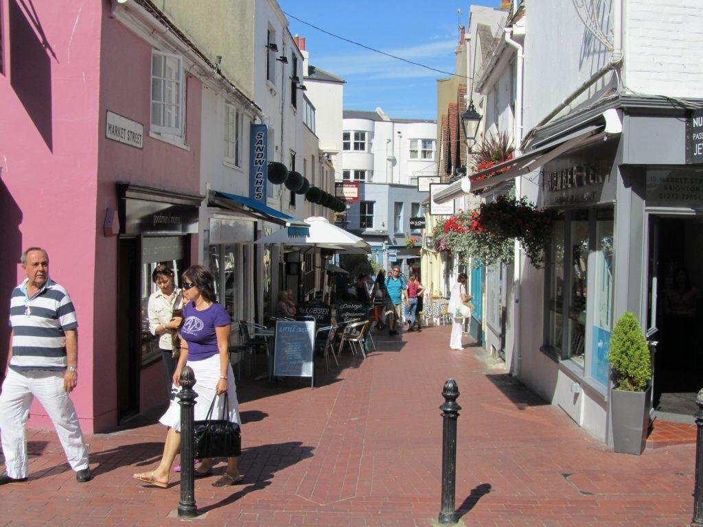 Narrow street named Market Street with people walking and sitting at outdoor café tables, lined with buildings painted in white and pastel colours under a clear blue sky