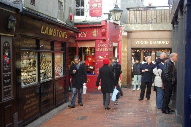 People walking and standing in a narrow shopping arcade with storefronts including Lambton's jewelry shop, The Lanes Armoury shop with a red facade, and Vogue Diamond store.