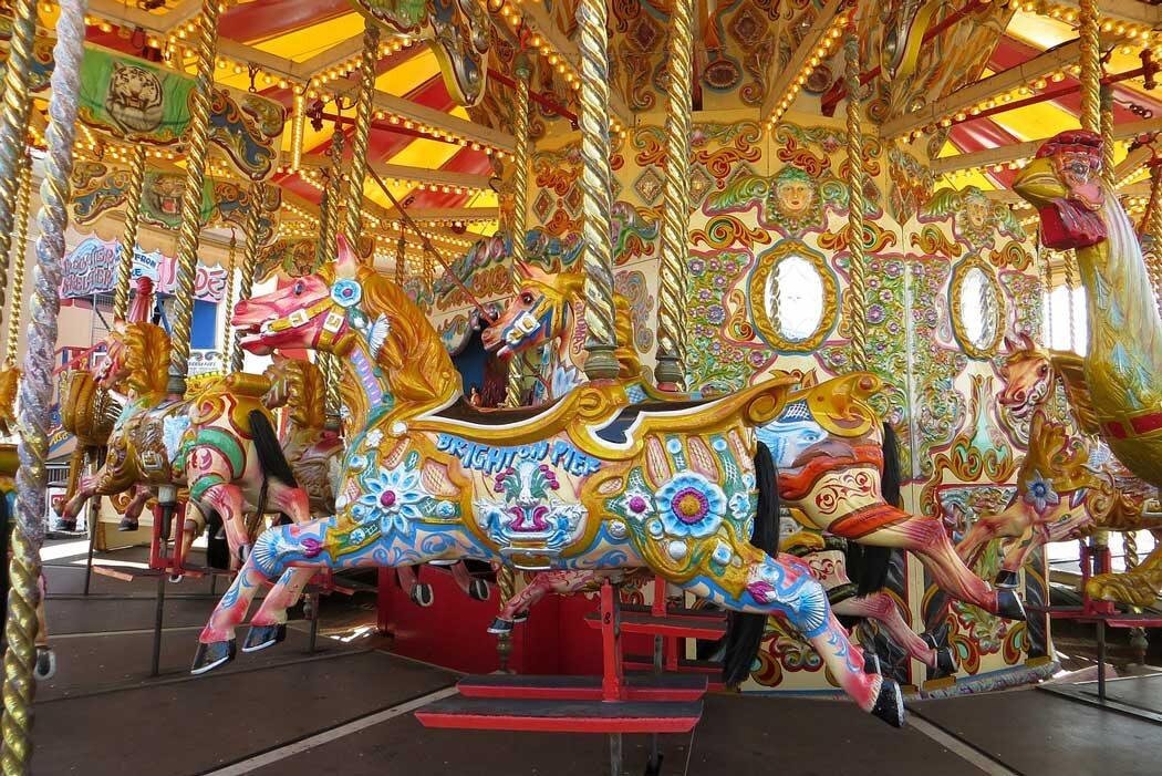 Colourfully decorated carousel horses with floral and ornamental designs, including a horse adorned with the words Brighton Pier, set on a brightly lit carousel with gold and red accents
