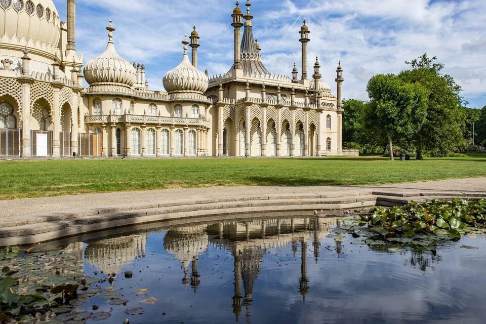 Bright historic building with ornate domes and spires reflected in a lily pond, set in a landscaped park with green grass and trees under a partly cloudy sky.
