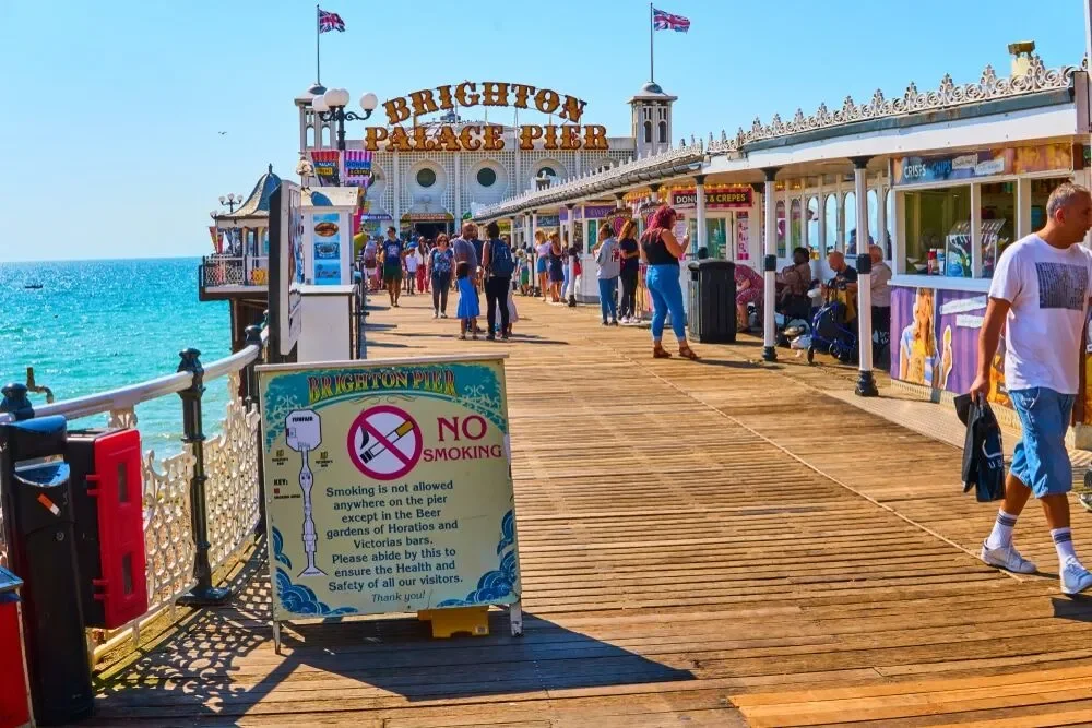 Brighton Palace Pier on a sunny day with people walking along the wooden boardwalk next to the sea, a large sign in the foreground stating no smoking allowed except in specific areas, and various shops and stalls on the right side under decorative railings and British flags.