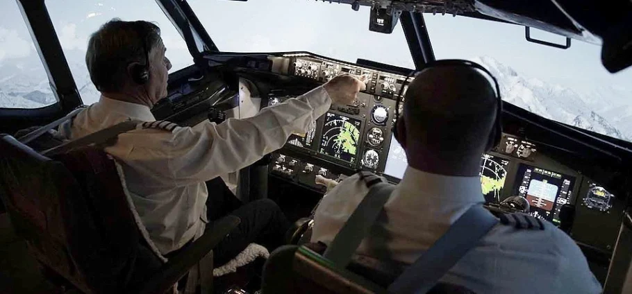 30 Minute Flight Simulator in Blackpool: Two uniformed pilots wearing headsets sit in a commercial aircraft cockpit, the left pilot reaching toward illuminated flight instruments and controls while snow-capped mountains are visible through the windshield.