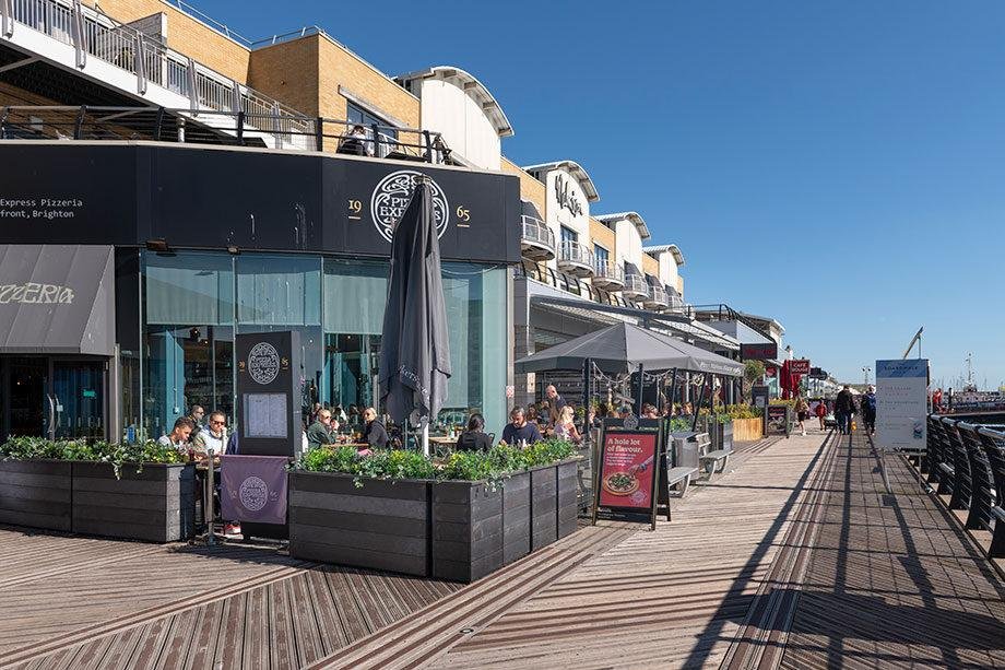 Outdoor seating area of a pizzeria on a wooden boardwalk with people dining, surrounded by planter boxes with greenery under a clear blue sky, with buildings and more cafes extending along the boardwalk by the sea.