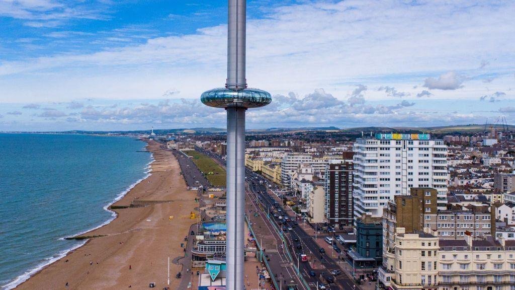Brighton i360: Aerial view of Brighton seafront showing the British Airways i360 observation tower next to the beach and a busy city road with buildings under a partly cloudy sky