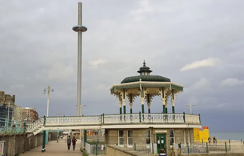 An ornate Victorian-style bandstand with a decorative roof and white railing on a promenade under a cloudy sky, with a tall observation tower and a few people walking nearby.