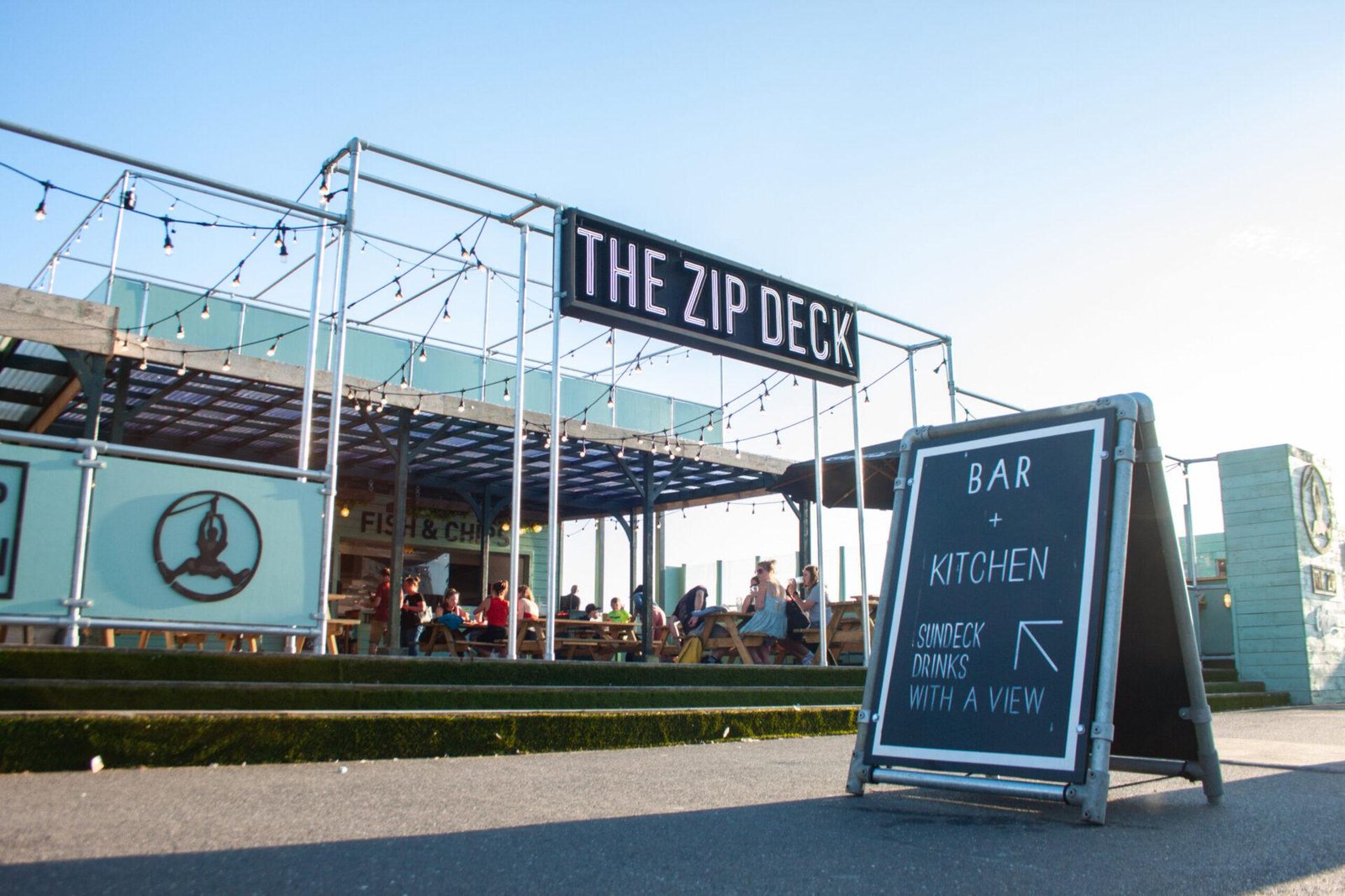 Outdoor seating area under a metal-framed structure with string lights and a sign reading 'THE ZIP DECK', people sitting at wooden picnic tables, and a sandwich board sign in the foreground advertising a bar and kitchen with sundeck drinks with a view.