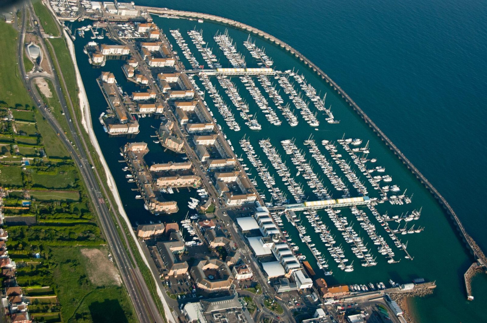 Brighton Marina: Aerial view of a marina with numerous boats docked in parallel rows, surrounded by residential buildings and roads on one side and open water on the other, with green fields and a highway visible nearby.