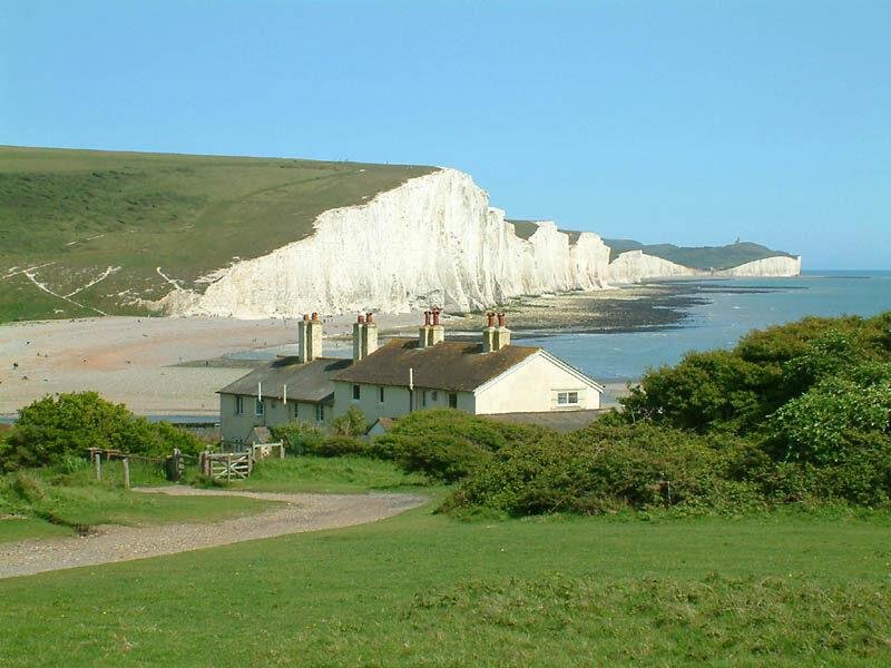 A coastal scene featuring white chalk cliffs extending into the distance along a calm shoreline, with a few houses and green grassy hills in the foreground under a clear blue sky.