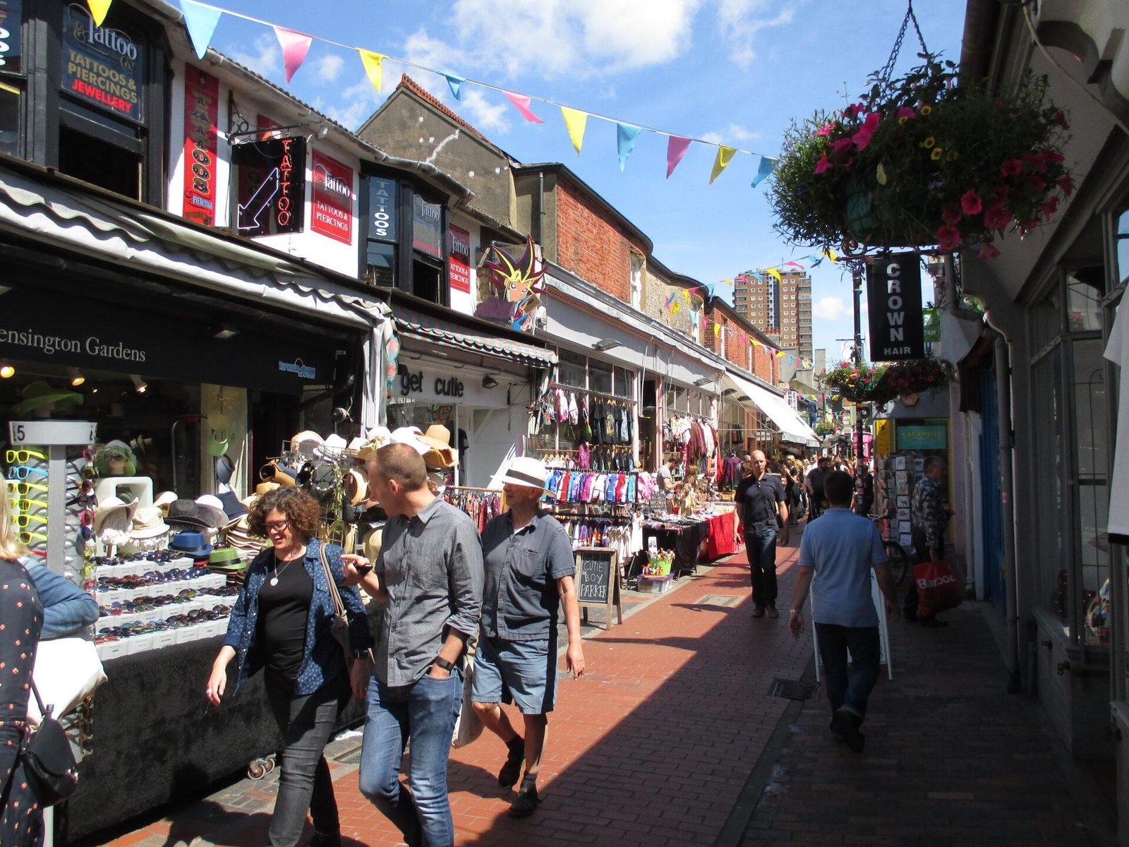 Busy outdoor market street with various shops, people walking along brick pavement, colourful bunting overhead, and hanging flower baskets, under a clear blue sky.
