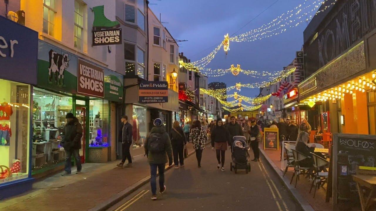 Crowded pedestrian street in the evening decorated with string lights overhead, lined with shops including a store named Vegetarian Shoes and a restaurant with outdoor seating on the right.