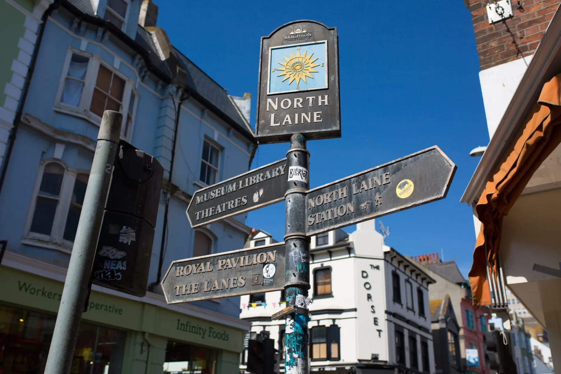 Metal directional signpost in a town centre with signs pointing to Museum Library Theatres, North Laine Station, Royal Pavilion The Lanes, and a top sign labeled North Laine under a blue sky, with Victorian buildings and shops including Infinity Foods and The Dorset in the background.