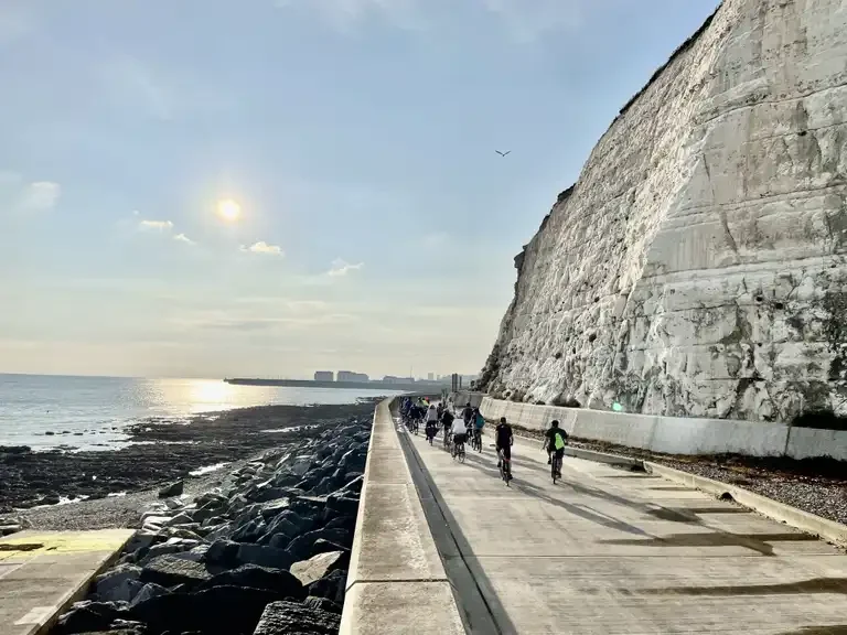 Undercliff Walk: Group of cyclists riding along a coastal pathway next to large white cliffs with the sun low in the sky over the sea