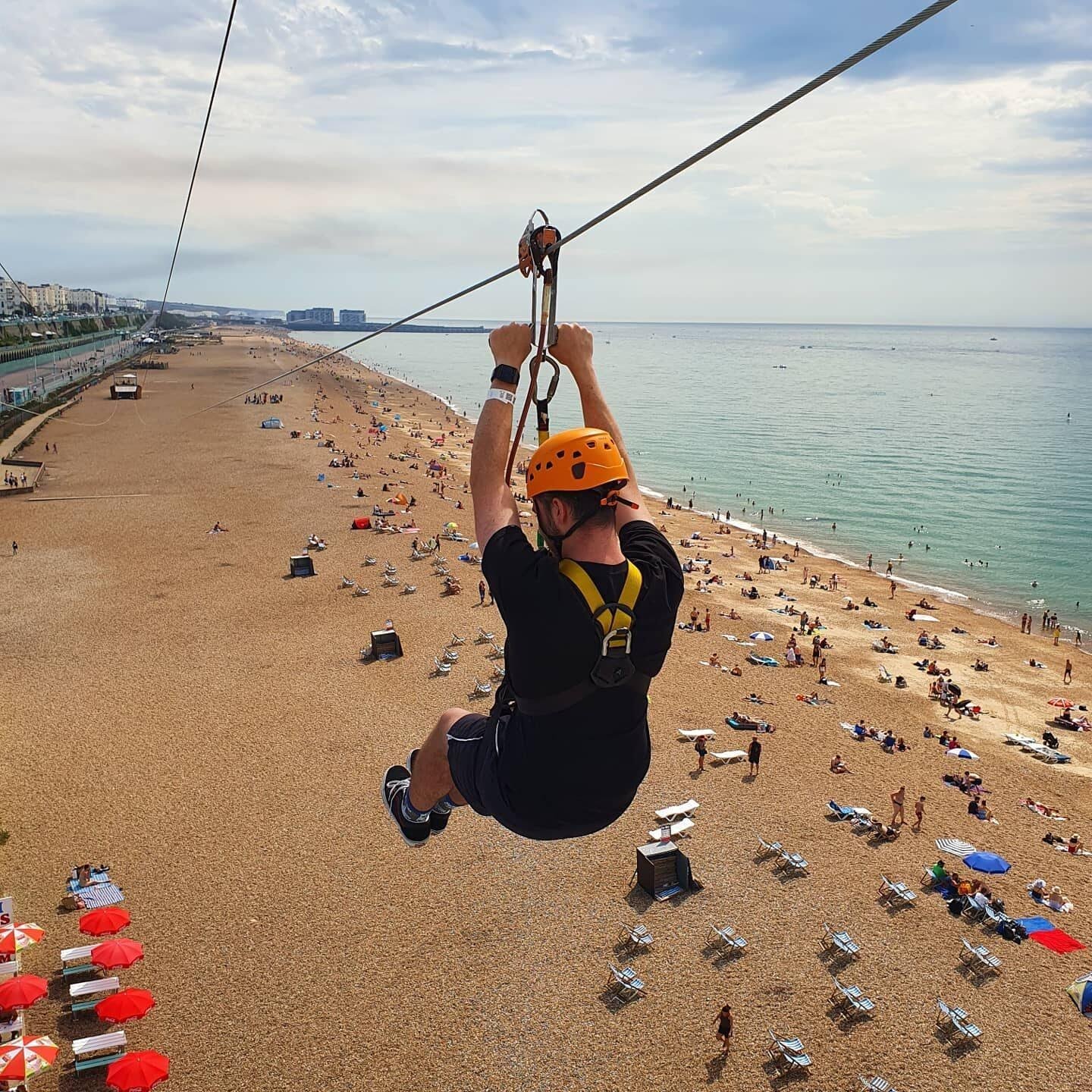 Person wearing an orange helmet and black clothing ziplining over a crowded beach with numerous people, umbrellas, and chairs scattered on the sand beside a calm sea under a partly cloudy sky.