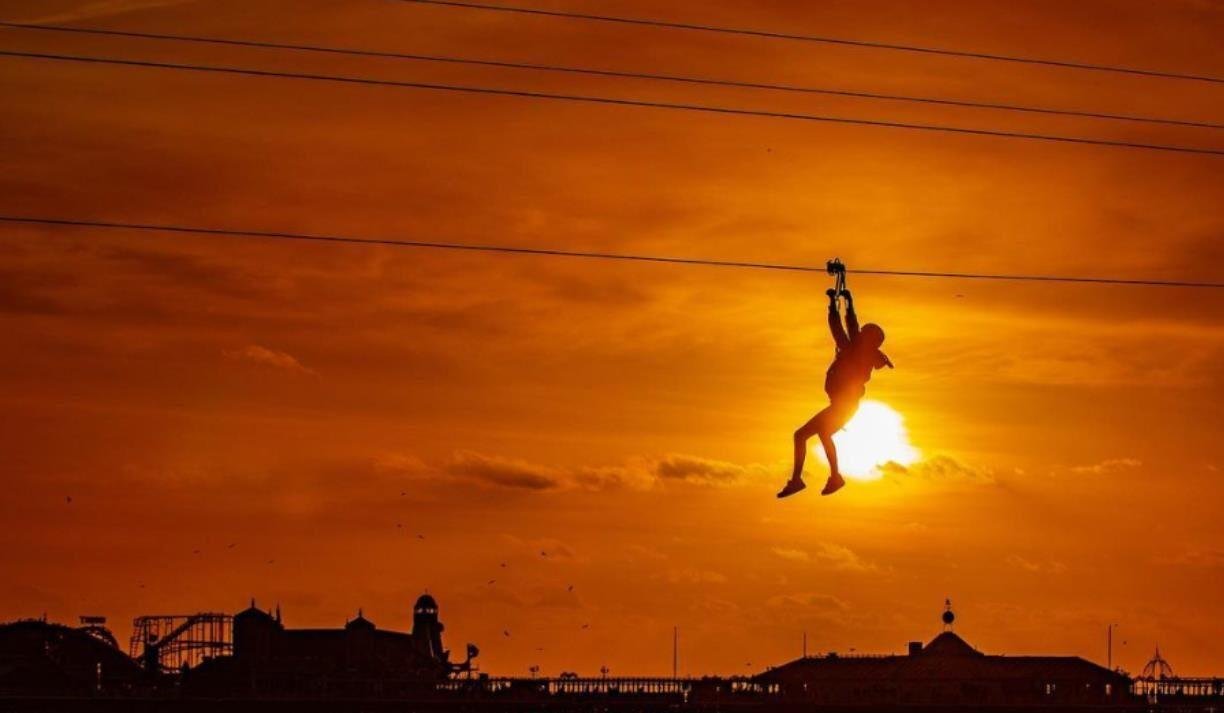 Silhouette of a person on a zip line against an orange sunset sky, with buildings and a roller coaster silhouette below.
