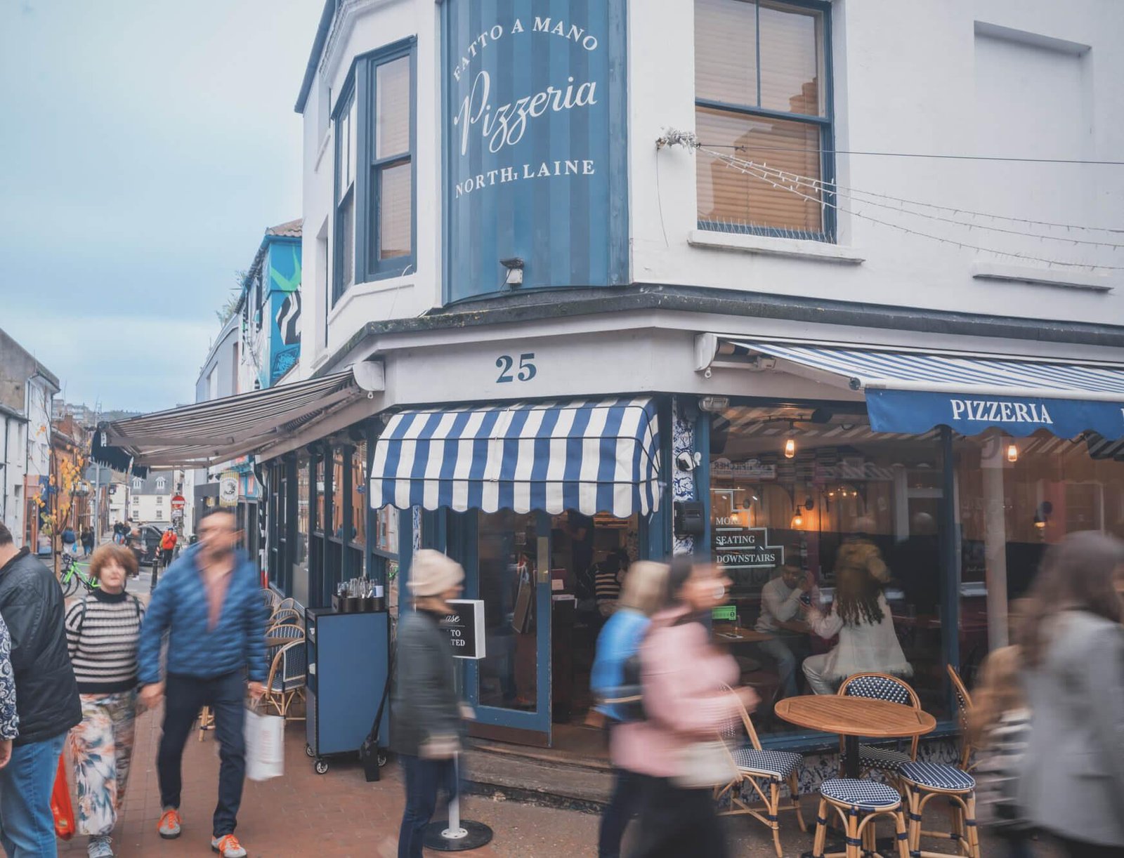Busy street scene outside Pizzeria North Laine at number 25, featuring a blue and white striped awning, outdoor seating with empty chairs and tables, and several blurred pedestrians walking by.