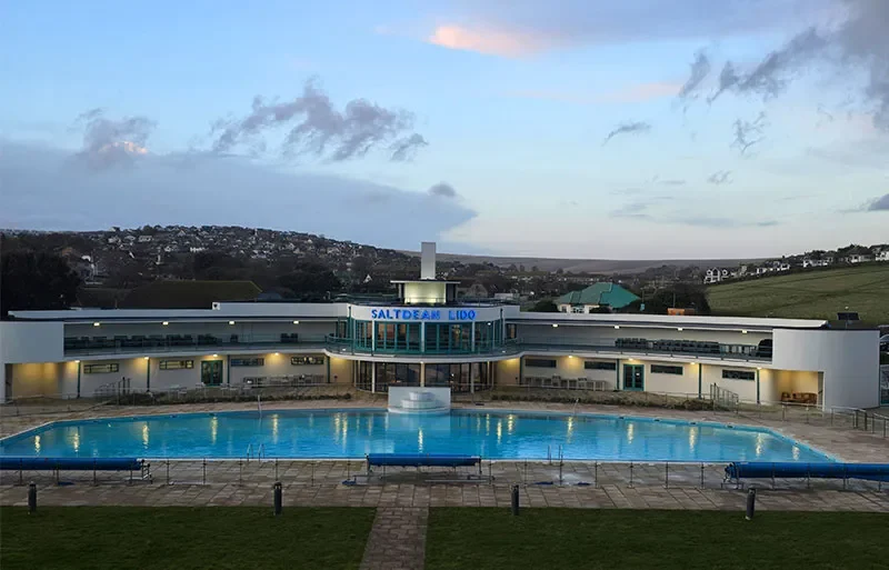 Saltdean Lido outdoor swimming pool and curved white building at dusk with hills and houses in the background