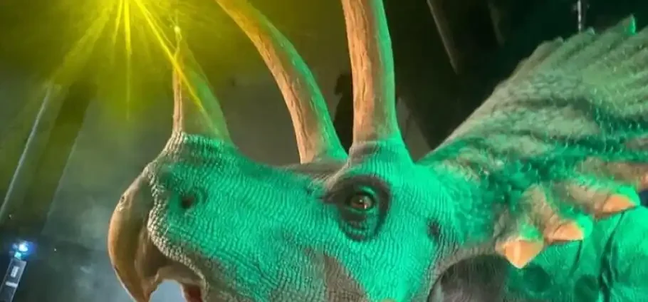 Blackpool Dino Explorers Adventure Experience: A Close-up of a life-sized green animatronic dinosaur head with three upward-curving horns, textured scaly skin, a visible eye and frilled neck with orange-tipped plates, bathed in green and yellow stage lighting