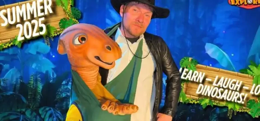 Man wearing a leather jacket and wide-brim hat poses with a friendly orange dinosaur puppet against a jungle-themed promotional backdrop displaying Summer 2025 and the slogan Learn - Laugh - Dinosaurs!