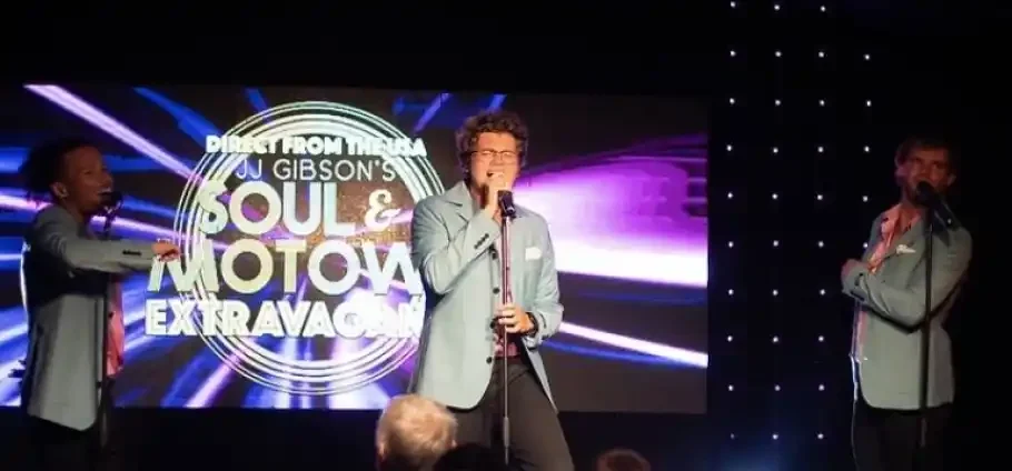 Three male vocalists in matching light-blue jackets and pink shirts perform on stage in front of a large illuminated backdrop reading 'Soul & Motown Extravaganza', the centre singer holding a microphone with purple light streaks and audience heads visible in the foreground.