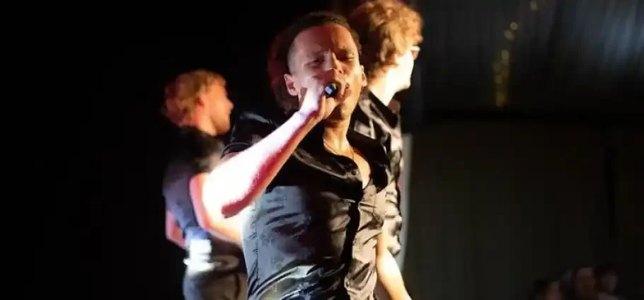 Male singer in a dark shirt singing passionately into a handheld microphone on a dimly lit stage, with two blurred backing performers and warm stage lighting.