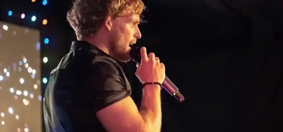Man with short curly blond hair in a black shirt holds a microphone to his mouth while performing on a dimly lit stage with out-of-focus coloured lights behind him.