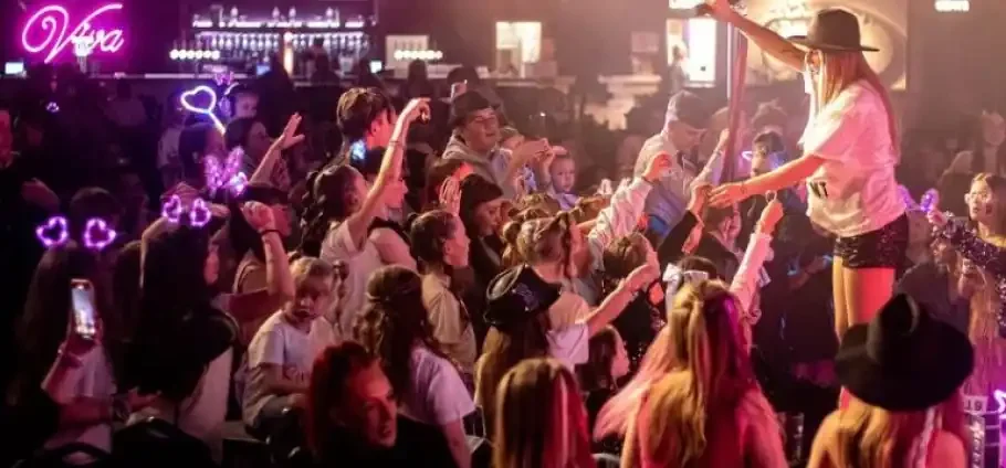 A female performer in a hat and shorts stands on stage under pink-purple lighting, reaching out to a packed crowd of mostly young fans who raise their hands and wear glowing heart-shaped headbands, with a neon "Viva" sign and bar visible in the background.