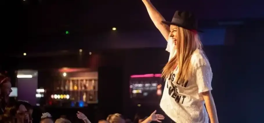 Blackpool Ultimate Pop Party Concert Experience: A Performer in a wide-brimmed hat and white T-shirt smiles with one arm raised while audience hands reach up in a dimly lit club with a bar visible in the background.