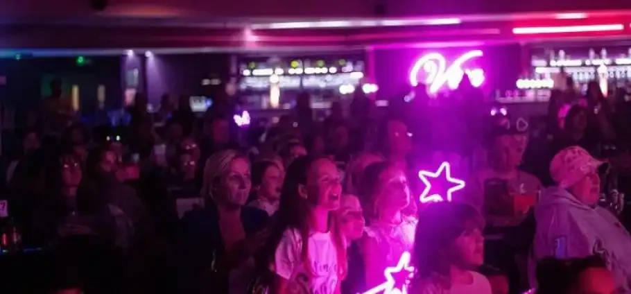 Crowd of people, including children, watching a performance in a dim indoor venue illuminated by pink and purple neon signs and handheld star-shaped light wands.