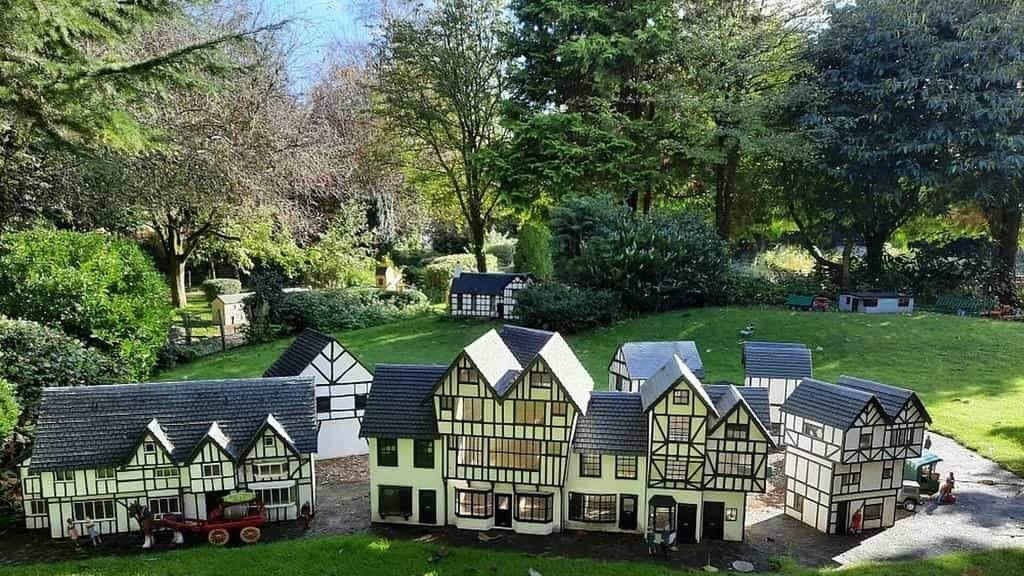 Blackpool Model Village and Gardens: A Cluster of miniature half‑timbered Tudor‑style model houses with black beams and cream walls arranged on a small paved area and lawn in a leafy garden, with trees in the background and tiny figures and a red cart visible.