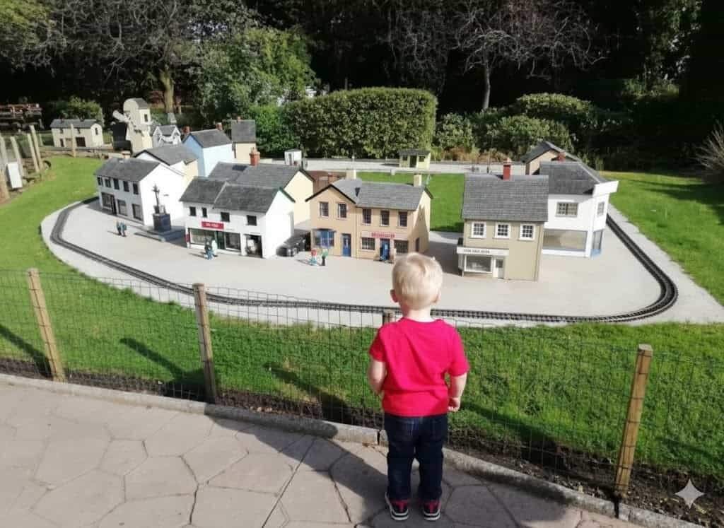 A small blond child in a red T-shirt stands with their back to the camera watching a detailed outdoor miniature village of model houses set inside an oval model railway behind a low wire fence.