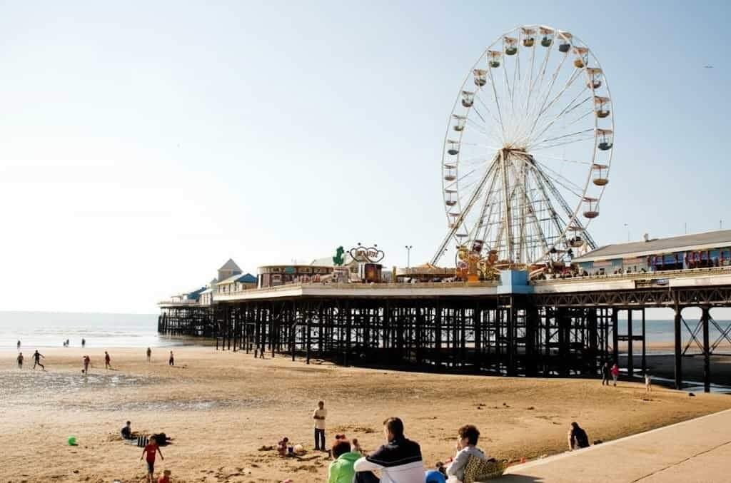 Seaside amusement pier with a large Ferris wheel and colourful cabins stretching over a sandy beach where people sit, walk and play on a sunny day.