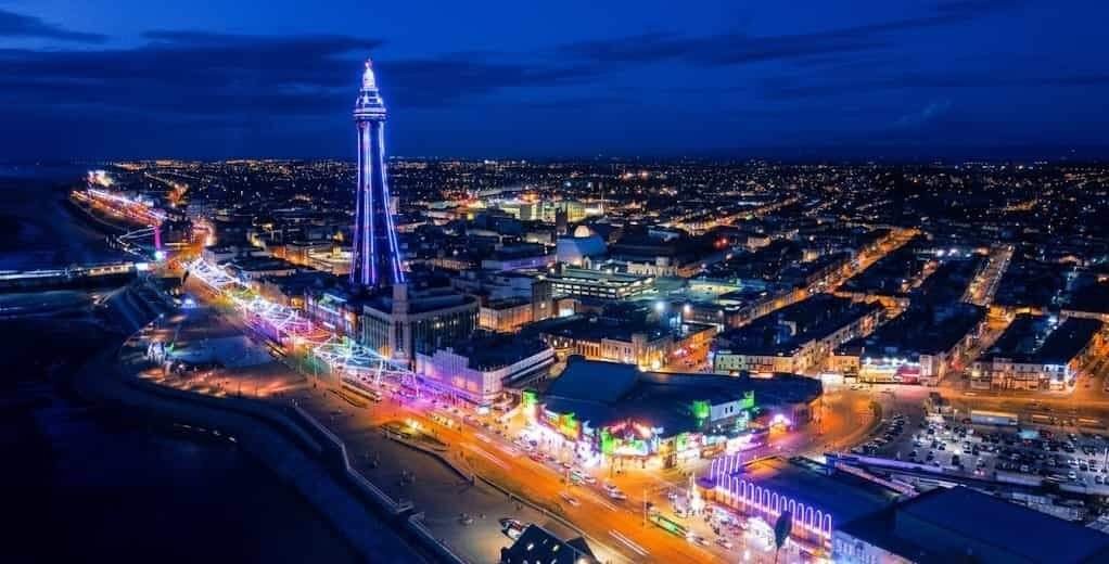 Aerial night view of Blackpool's illuminated seafront with the tall, lit-up Blackpool Tower rising above a colourful neon-lit promenade and amusements, the dark coastline to the left and city lights stretching to the horizon.