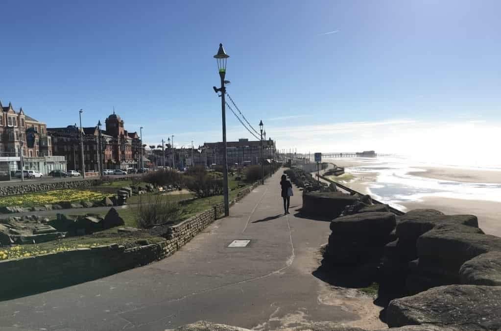 Blackpool Seafront: A Sunny coastal promenade with a lone walker heading toward a distant pier, Victorian seaside buildings and daffodil-filled gardens on the left and sandy beach and rocks on the right.