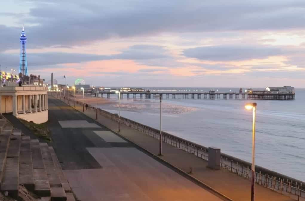 Seafront promenade at dusk with a lit observation tower and Ferris wheel to the left, a long pier stretching into the calm sea under a pastel pink-and-blue sky, and a mostly empty walkway lit by street lamps.