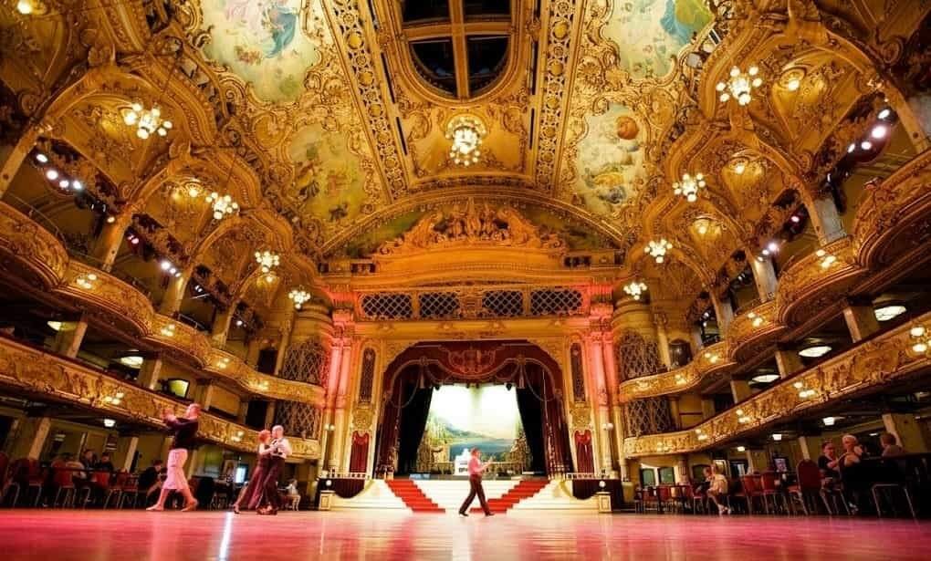 Ornately decorated golden ballroom with a painted vaulted ceiling, crystal chandeliers, tiered balconies and a central stage with red‑carpeted steps while couples dance on the polished floor.