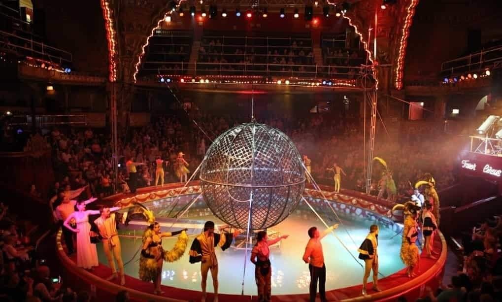 Circus ring illuminated by warm spotlights with a large metal spherical stunt cage at the centre, surrounded by costumed performers taking a bow and a packed audience in the balconies.