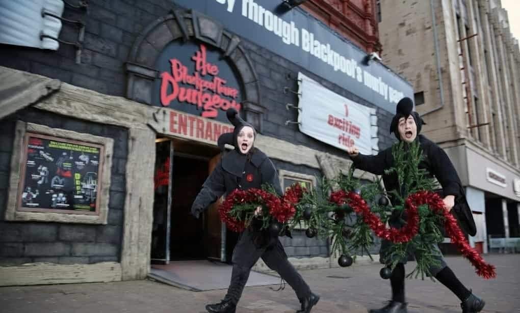 Two performers in black Victorian-style costumes with surprised expressions carry a large festive garland trimmed with red tinsel and black baubles past the entrance to The Blackpool Tower Dungeon on a city street.