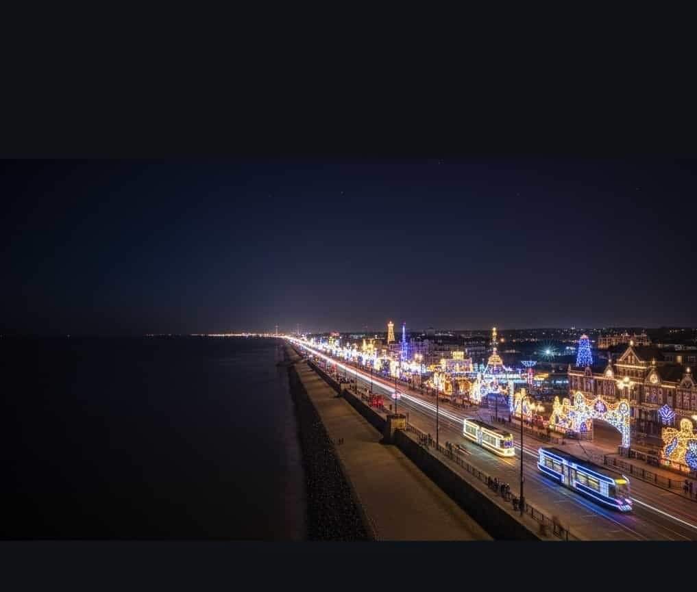 Long-exposure view of a night-time seafront boulevard with two trams streaking past ornate festive light displays and Victorian-style buildings on the right and a dark, calm sea on the left.