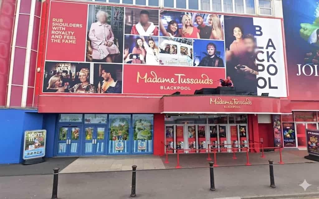Madame Tussauds Blackpool façade with a large red billboard showing a collage of celebrity portraits, a red canopy over multiple entrance doors with blue door frames, and queue barriers and short black bollards on the pavement.