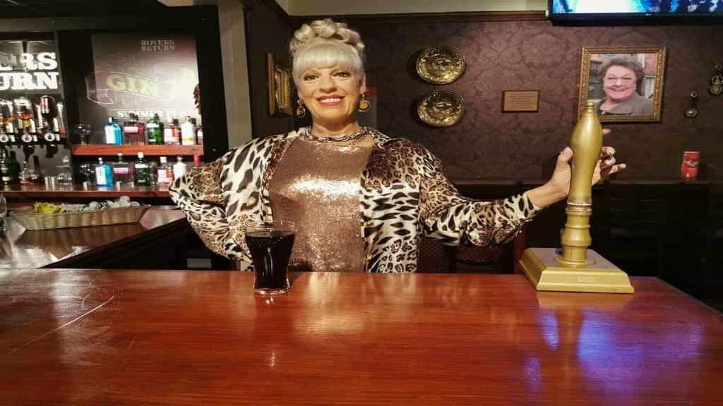 Smiling woman with a blonde updo wearing a leopard-print jacket and sequinned top stands behind a pub bar, one hand on a brass beer pump and a pint glass on the counter in front of her.