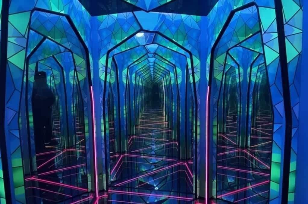 Ripley's Believe It or Not! Blackpool: Mirrored installation forming an endless blue-green geometric arched corridor with pink neon floor lines and repeating reflections, a dark human silhouette visible at the left.