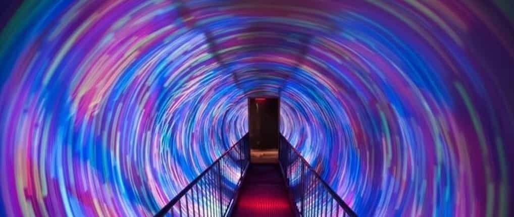 A narrow metal-railed walkway leads into a circular tunnel lined with swirling blue, purple and pink light patterns, ending at a dark doorway.