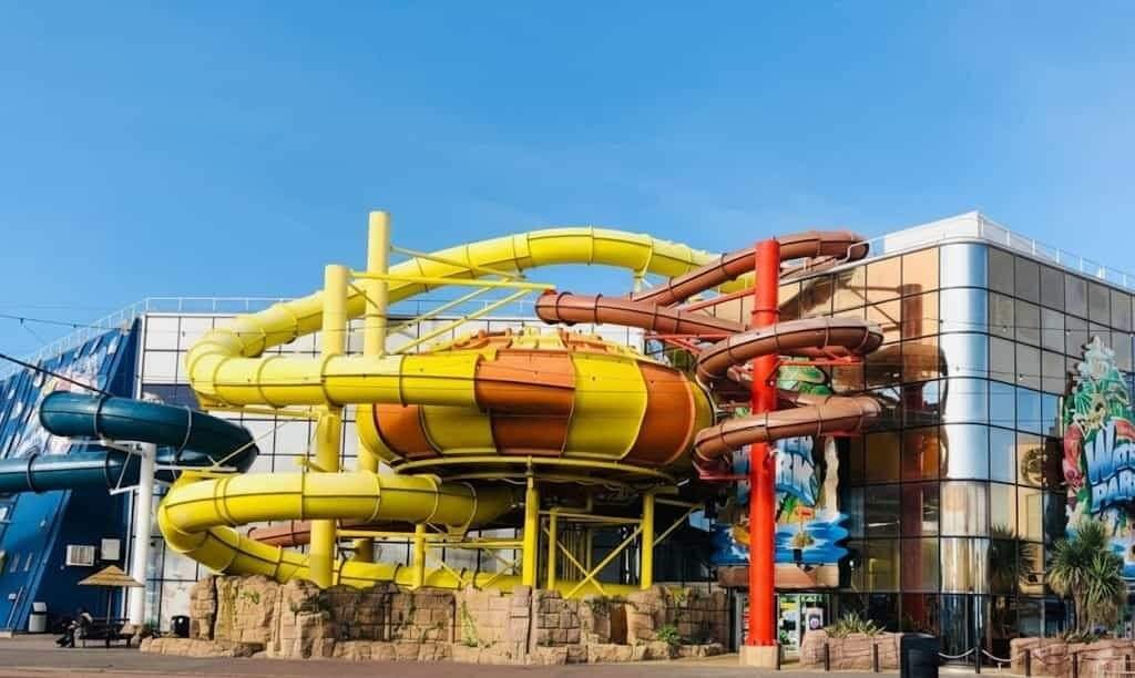 Exterior of a water park featuring bright yellow tubular slides and a brown spiral slide winding into a large orange-and-yellow bowl, supported by red pillars and anchored on faux rock at the base of a mirrored-glass building under a clear blue sky.