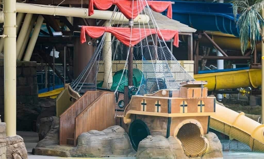 Toy pirate-ship play structure with red fabric sails and rigging over a wooden deck and rock-like base, featuring netted climbing areas, a round tunnel entrance and yellow tube slides in an indoor waterpark.
