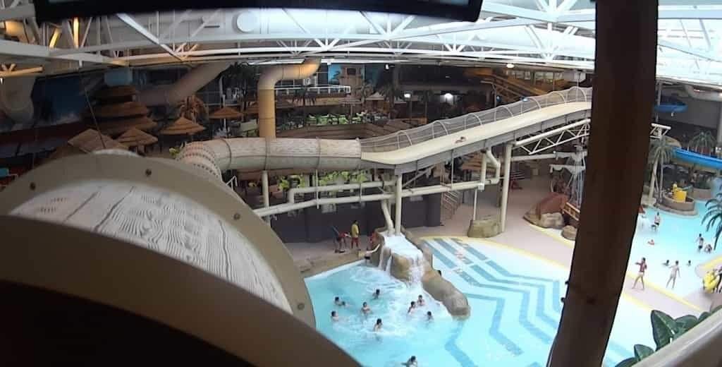 Sandcastle Waterpark Blackpool: A High-angle view of a busy indoor waterpark with enclosed tube slides and elevated walkways above a wave pool where people swim and wade around artificial rocks and palm-tree decorations beneath a white steel roof