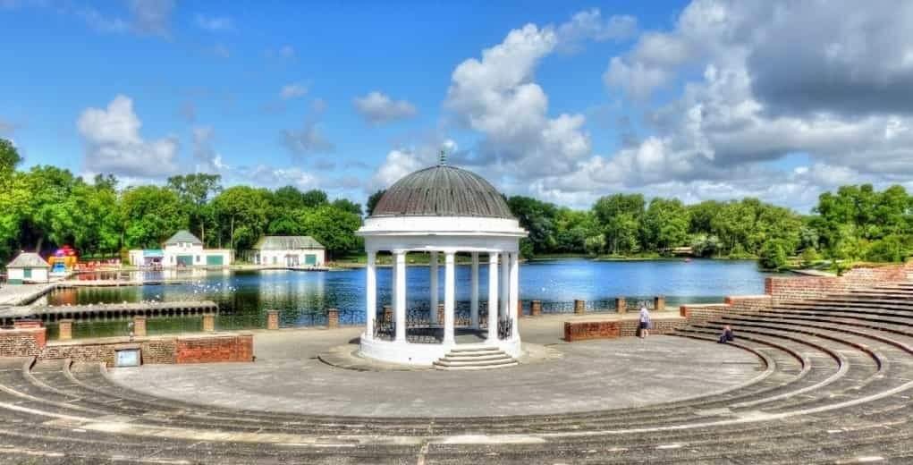 White domed pavilion with classical columns stands at the centre of a semicircular stone amphitheatre overlooking a calm lake lined with green trees and small boathouses under a bright blue sky with scattered clouds.